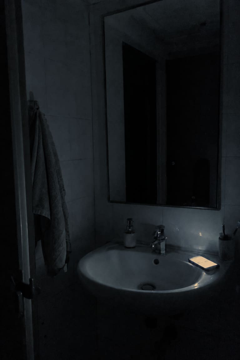 A small bathroom at night with the lights off: a dark mirror above a white sink.